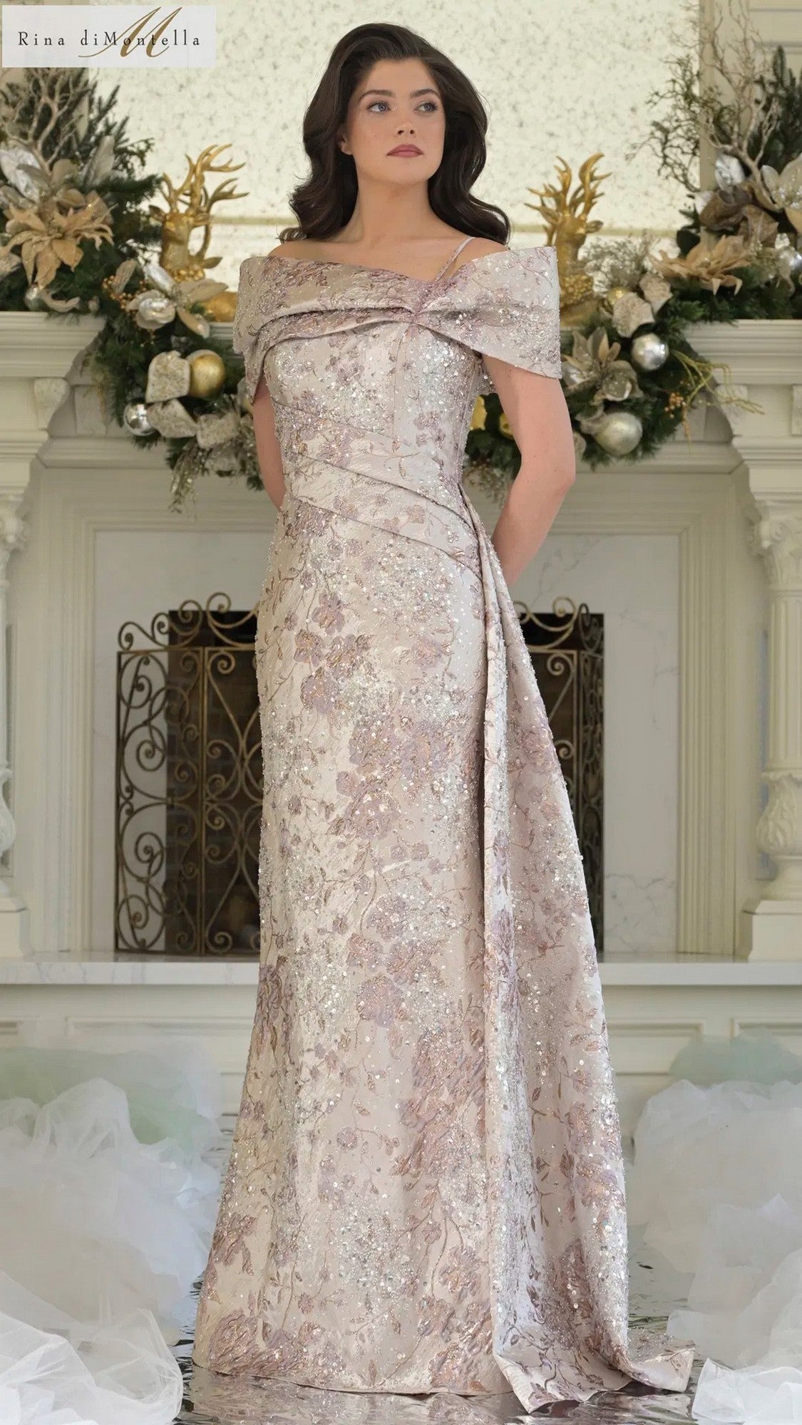 A woman stands indoors wearing the Rina Di Montella RD3306 long formal dress in Rose Multi, featuring off-the-shoulder jacquard fabric with shimmering metallic floral details. Ornate holiday decorations are displayed on the mantel behind her.